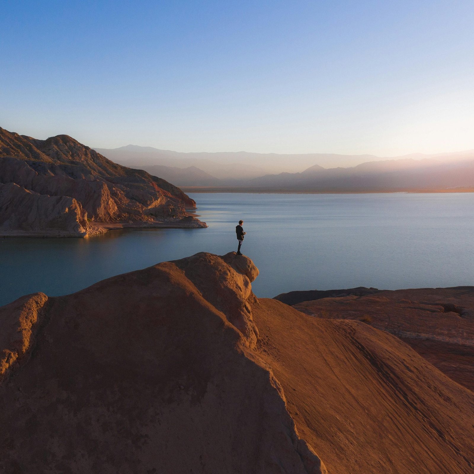 A lone individual stands on a rocky peak overlooking a tranquil lake at sunrise.