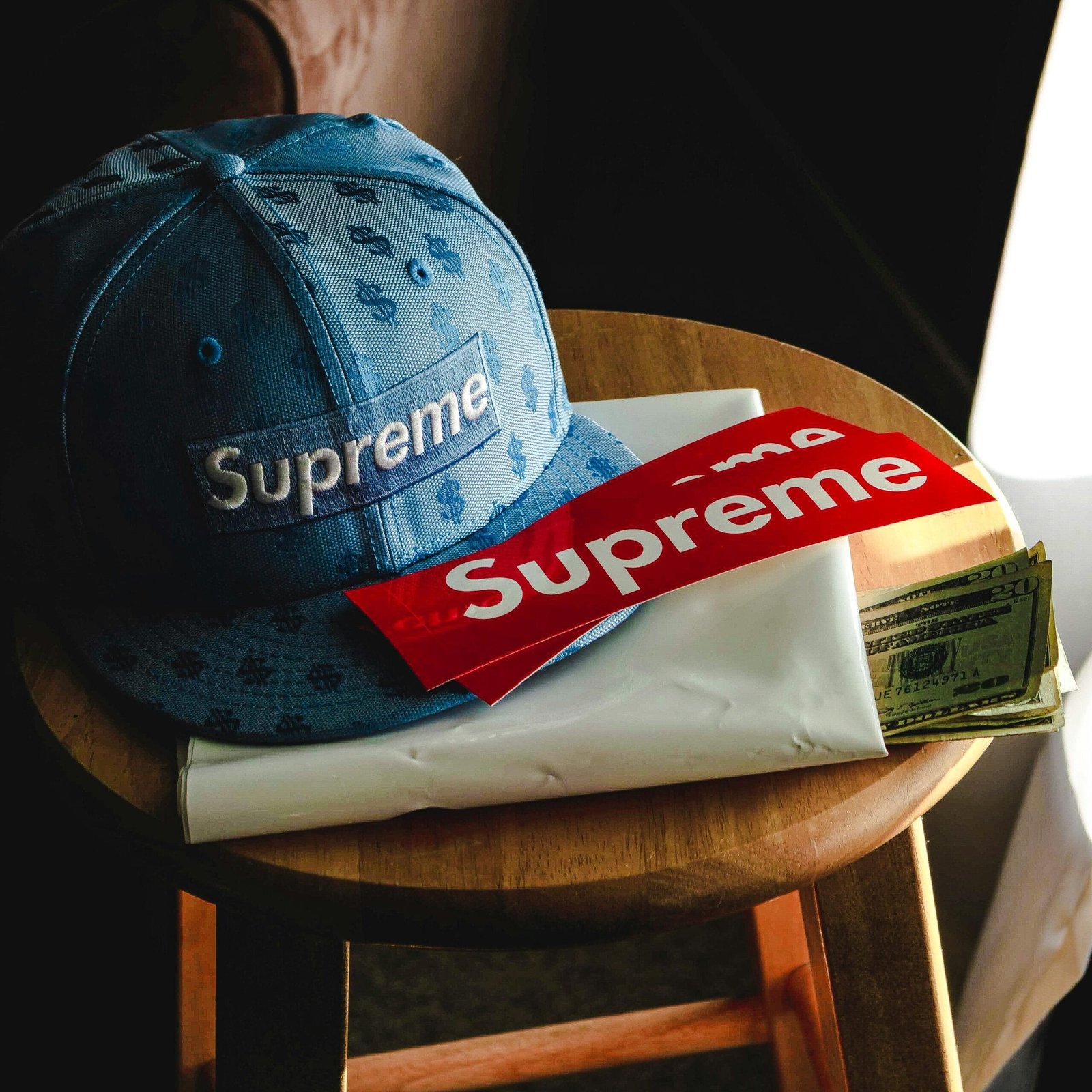 Close-up of a Supreme cap, banknotes, and stickers on a wooden stool indoors.