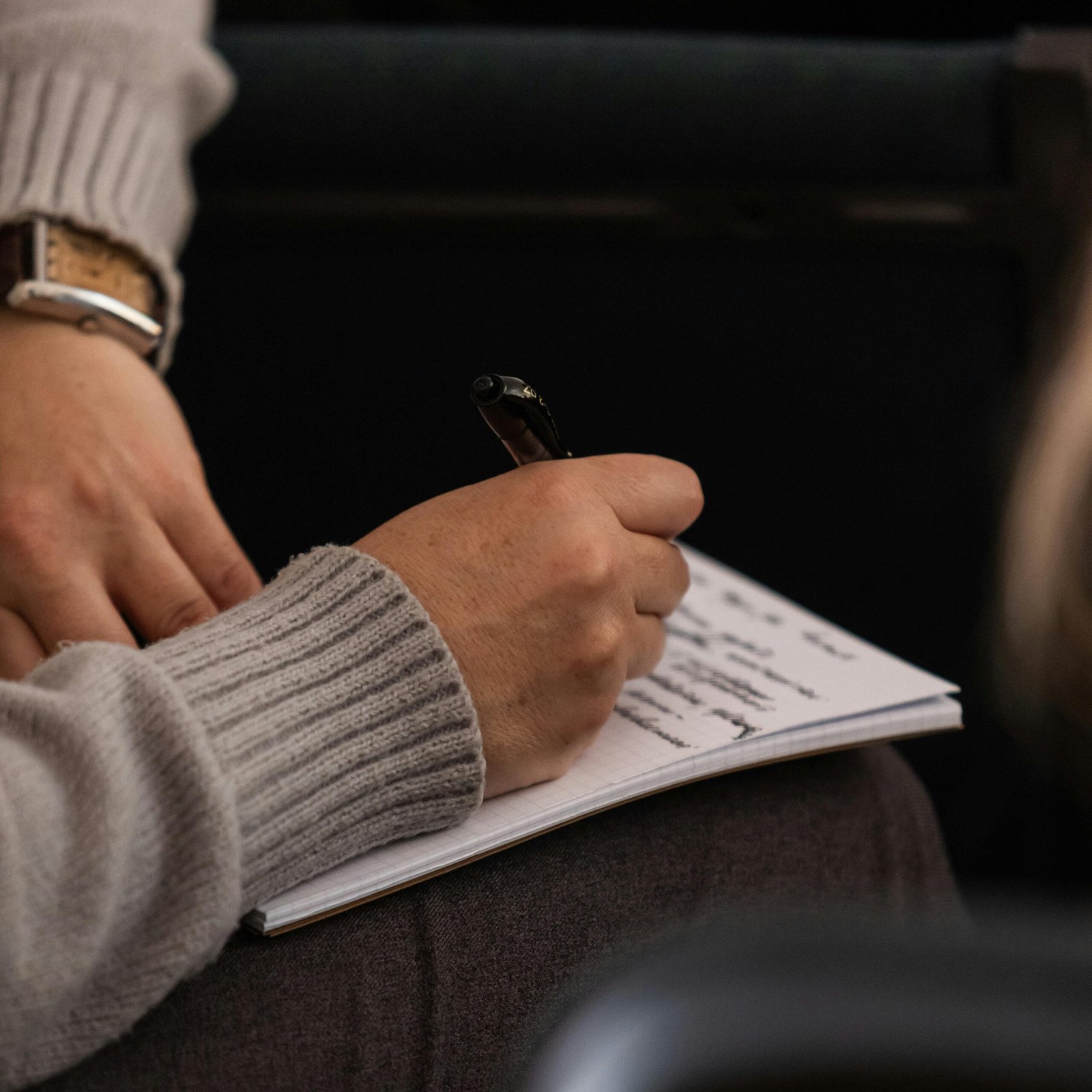 Person in grey sweater taking notes in a notebook with a pen.