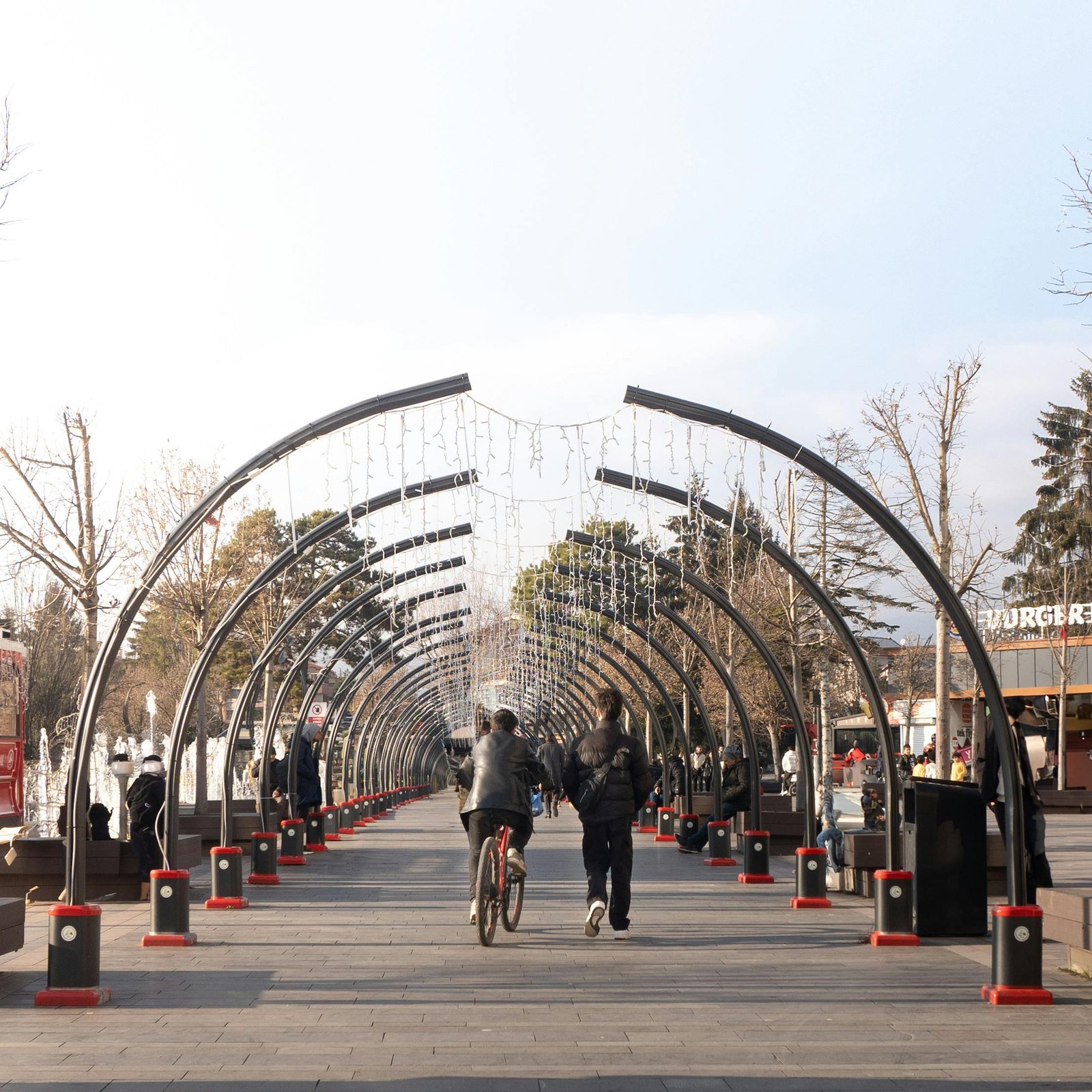 A promenade with an arched walkway and people strolling and cycling on a sunny day.