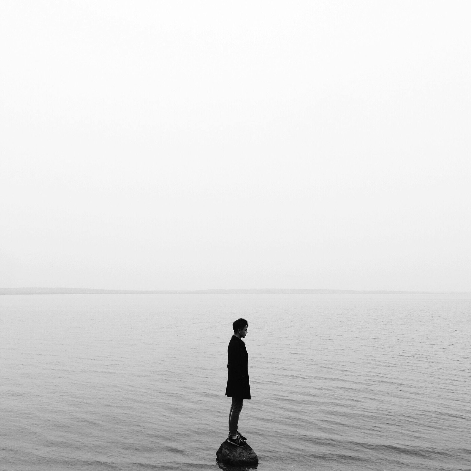 Black and white image of a solitary person on a rock in a calm lake, evoking solitude and reflection.