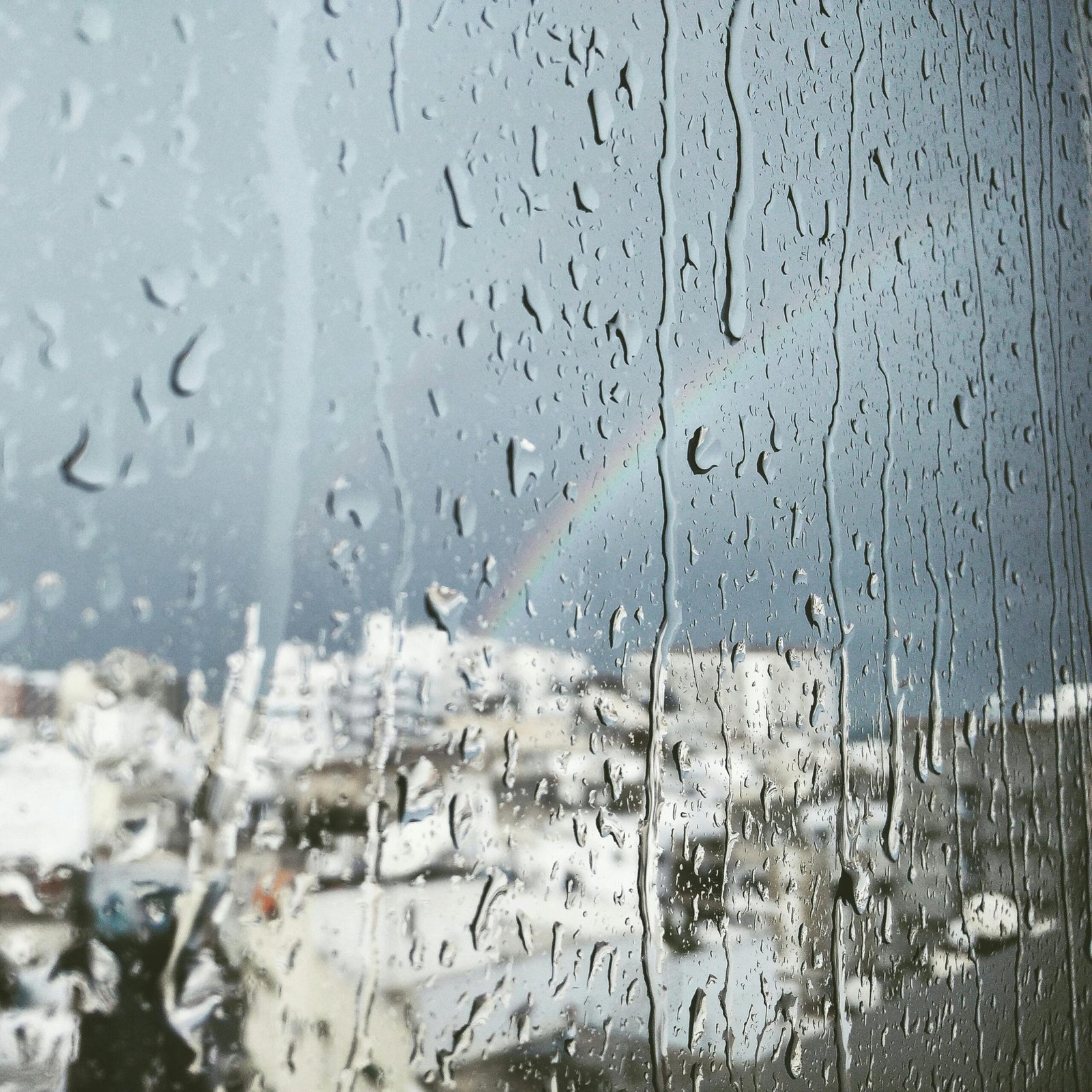 Raindrops on glass with a faint rainbow and cityscape view in the background.