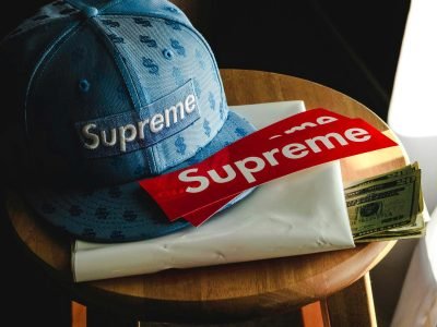 Close-up of a Supreme cap, banknotes, and stickers on a wooden stool indoors.