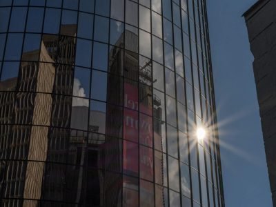 Glass skyscraper reflecting cityscape with sunburst in urban setting.