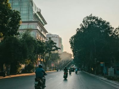 Motorcyclists commute on a tree-lined street near PVcomBank building at sunrise.