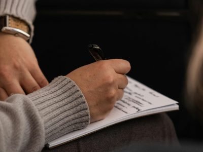 Person in grey sweater taking notes in a notebook with a pen.