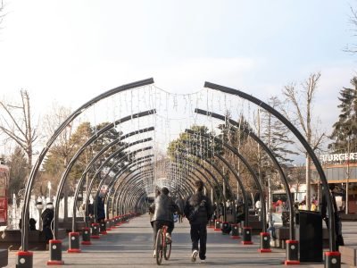 A promenade with an arched walkway and people strolling and cycling on a sunny day.
