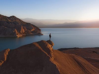 A lone individual stands on a rocky peak overlooking a tranquil lake at sunrise.
