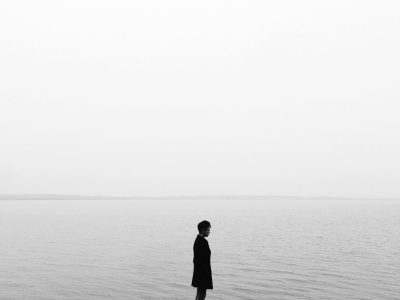 Black and white image of a solitary person on a rock in a calm lake, evoking solitude and reflection.