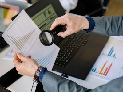 Businessman examines data charts with a magnifying glass over a laptop in an office setting.