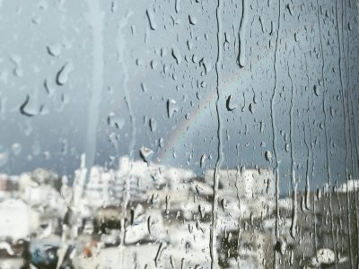 Raindrops on glass with a faint rainbow and cityscape view in the background.